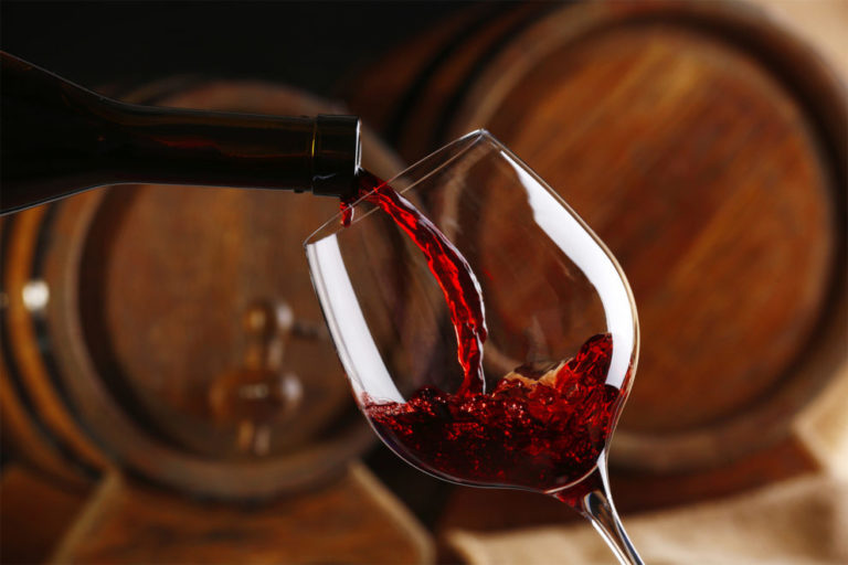 Pouring red wine from bottle into glass with wooden wine casks o